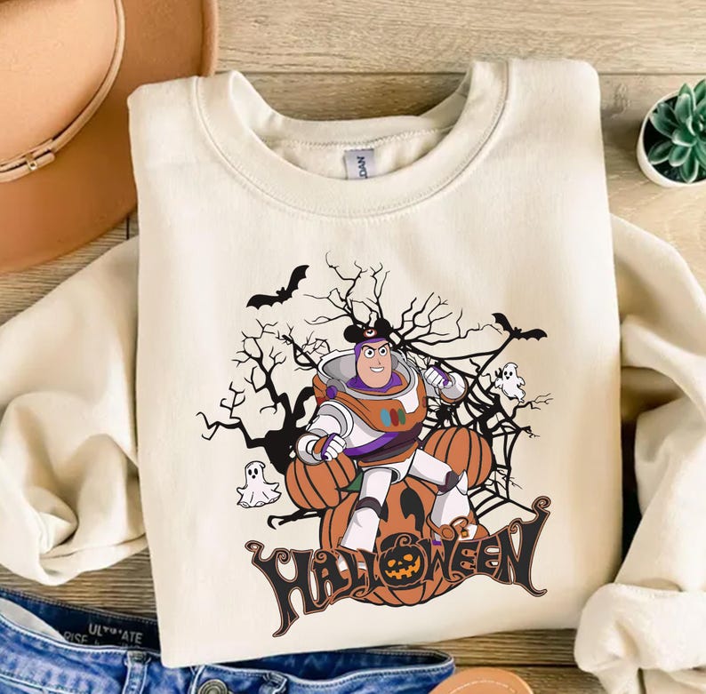K&ouml;nnte beinhalten: Cremefarbenes Sweatshirt mit einem Halloween-Motiv. Das Design zeigt eine Zeichentrickfigur in einem Raumanzug, K&uuml;rbisse, Flederm&auml;use, Geister und das Wort "HALLOWEEN". Die Grafik ist in Orange, Schwarz, Wei&szlig; und Lila gehalten.