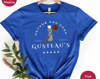 May include: A royal blue t-shirt with a white graphic of a rat chef holding a wooden spoon. The graphic says "Anyone Can Cook" with "Paris" and "France" on either side of the spoon. The text "Gusteau's" is below the graphic with five white stars.