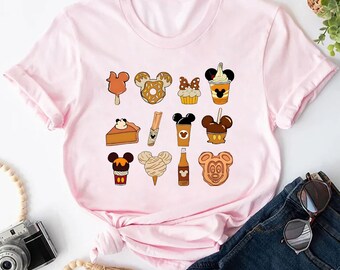 May include: A light pink t-shirt featuring a variety of cartoon food illustrations. The illustrations include ice cream, donuts, cupcakes, and other treats, all in a cartoon style. The shirt is made of a soft material.