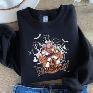K&ouml;nnte beinhalten: Schwarzes Sweatshirt mit einem Halloween-Motiv. Das Design zeigt eine Zeichentrickfigur in einem Raumanzug, K&uuml;rbisse, Flederm&auml;use und das Wort "HALLOWEEN". Das Sweatshirt besteht aus einem weichen Material.