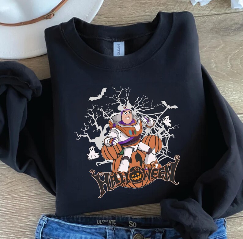 K&ouml;nnte beinhalten: Schwarzes Sweatshirt mit einem Halloween-Motiv. Das Design zeigt eine Zeichentrickfigur in einem Raumanzug, K&uuml;rbisse, Flederm&auml;use und das Wort "HALLOWEEN". Das Sweatshirt besteht aus einem weichen Material.