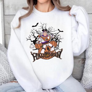 K&ouml;nnte beinhalten: Wei&szlig;es Sweatshirt mit einem Halloween-Motiv. Das Design zeigt eine Zeichentrickfigur, K&uuml;rbisse, Flederm&auml;use und das Wort "Halloween". Die Grafik ist in Orange, Lila und Schwarz gehalten.