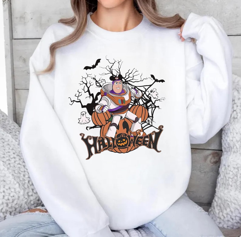 K&ouml;nnte beinhalten: Wei&szlig;es Sweatshirt mit einem Halloween-Motiv. Das Design zeigt eine Zeichentrickfigur, K&uuml;rbisse, Flederm&auml;use und das Wort "Halloween". Die Grafik ist in Orange, Lila und Schwarz gehalten.