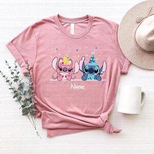 May include: Pink and blue Stitch characters wearing party hats with confetti in the background. The shirt is pink and has the word "Name" printed below the characters.