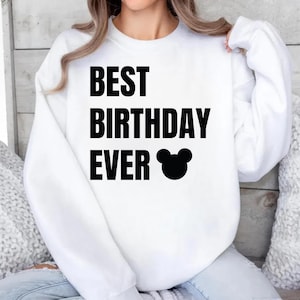 May include: White sweatshirt with the text "BEST BIRTHDAY EVER" in bold black letters, accompanied by a black Mickey Mouse head silhouette. The long-sleeved top is a casual style.