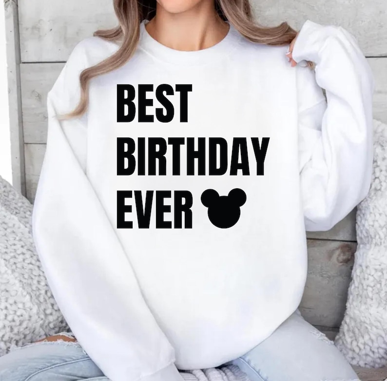 May include: White sweatshirt with the text "BEST BIRTHDAY EVER" in bold black letters, accompanied by a black Mickey Mouse head silhouette. The long-sleeved top is a casual style.