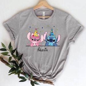 May include: Gray t-shirt with a cartoon image of Stitch and Angel from Lilo & Stitch wearing party hats. The text "Alexis" is printed below the characters.