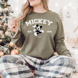 May include: Olive green sweatshirt featuring the word "MICKEY" in white block letters above a black and white graphic of Mickey Mouse. The text "EST. 1928" is also displayed. The sweatshirt is part of a casual clothing collection.