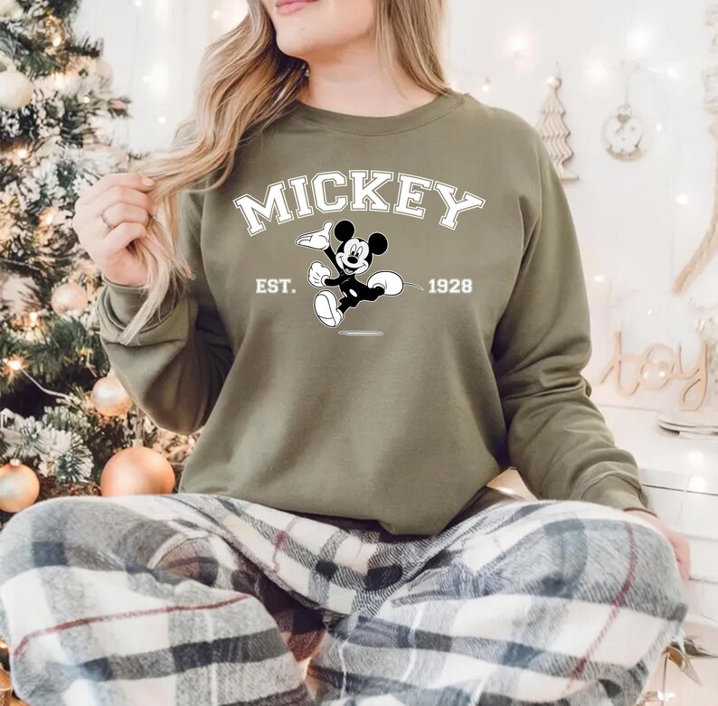May include: Olive green sweatshirt featuring the word "MICKEY" in white block letters above a black and white graphic of Mickey Mouse. The text "EST. 1928" is also displayed. The sweatshirt is part of a casual clothing collection.