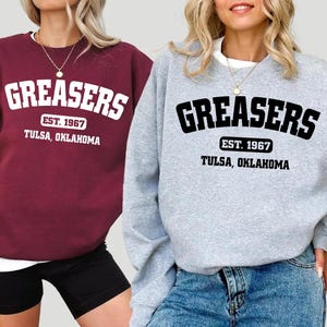 May include: Two women wearing gray and burgundy sweatshirts with the text "Greasers Est. 1967 Tulsa, Oklahoma" printed on the front.