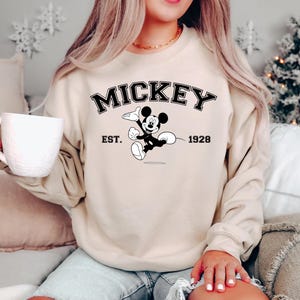 May include: Beige sweatshirt featuring the word "MICKEY" in black, with a graphic of Mickey Mouse and the text "EST. 1928". The sweatshirt is a crew neck style and has long sleeves. The overall theme is Disney.