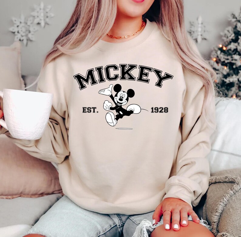 May include: Beige sweatshirt featuring the word "MICKEY" in black, with a graphic of Mickey Mouse and the text "EST. 1928". The sweatshirt is a crew neck style and has long sleeves. The overall theme is Disney.