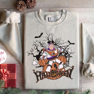 K&ouml;nnte beinhalten: Cremefarbenes Sweatshirt mit einem Halloween-Motiv. Das Design zeigt eine Zeichentrickfigur in einem Raumanzug, K&uuml;rbisse, Flederm&auml;use, Geister und das Wort "Halloween". Die Grafik ist in Orange, Schwarz und Wei&szlig; gehalten.