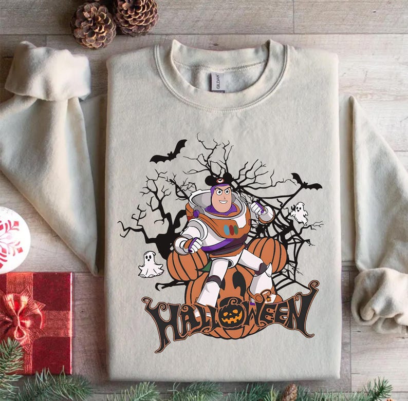 K&ouml;nnte beinhalten: Cremefarbenes Sweatshirt mit einem Halloween-Motiv. Das Design zeigt eine Zeichentrickfigur in einem Raumanzug, K&uuml;rbisse, Flederm&auml;use, Geister und das Wort "Halloween". Die Grafik ist in Orange, Schwarz und Wei&szlig; gehalten.