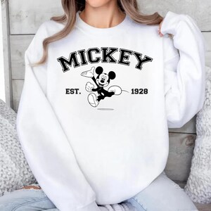 May include: White sweatshirt featuring a black and white graphic of Mickey Mouse. The word "MICKEY" is arched across the top, with "EST. 1928" below the graphic. The sweatshirt has long sleeves and a crew neck.
