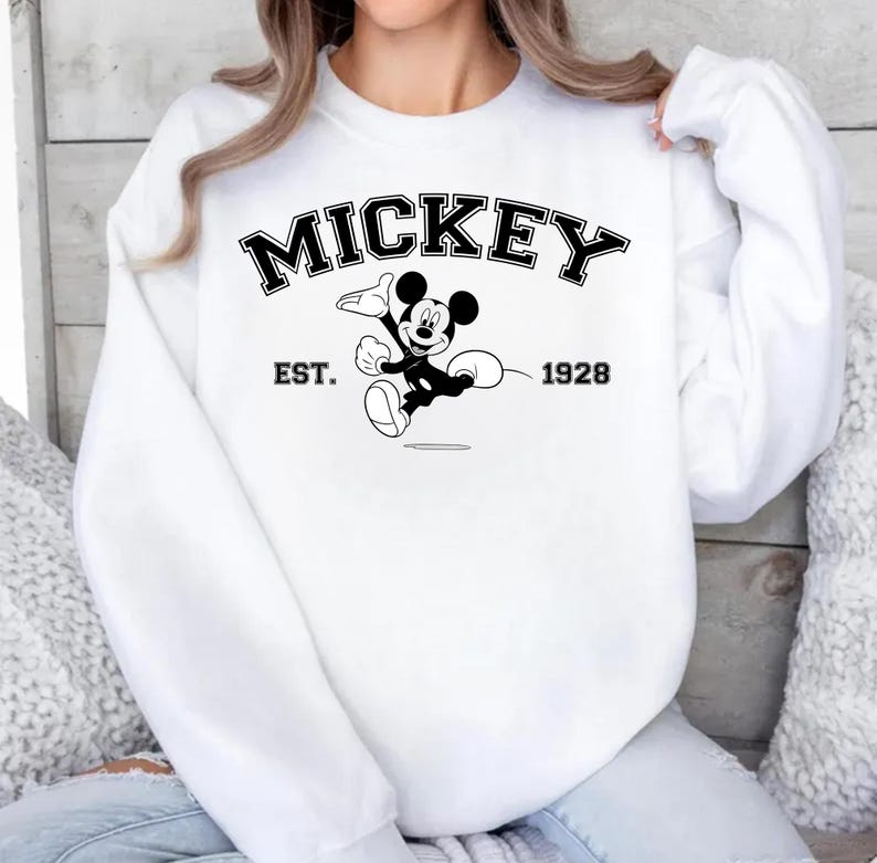 May include: White sweatshirt featuring a black and white graphic of Mickey Mouse. The word "MICKEY" is arched across the top, with "EST. 1928" below the graphic. The sweatshirt has long sleeves and a crew neck.