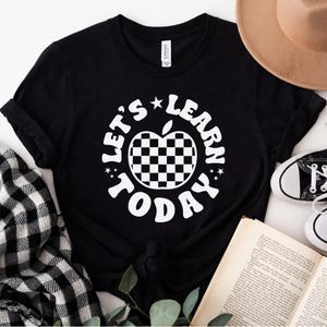 May include: Black t-shirt with the text "LET'S LEARN TODAY" in a white, circular design. The design includes a checkered apple graphic. A tan hat, black and white shoes, and a book are in the background.