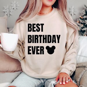 May include: A beige sweatshirt with the text "BEST BIRTHDAY EVER" in bold black letters, along with a black Mickey Mouse head graphic. The sweatshirt is a neutral color and has long sleeves.