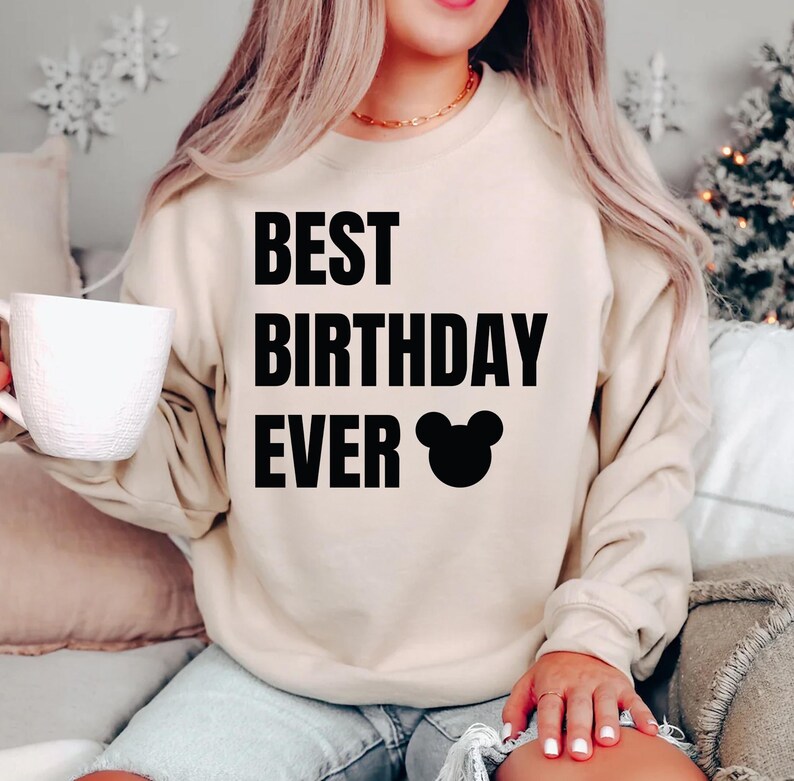 May include: A beige sweatshirt with the text "BEST BIRTHDAY EVER" in bold black letters, along with a black Mickey Mouse head graphic. The sweatshirt is a neutral color and has long sleeves.