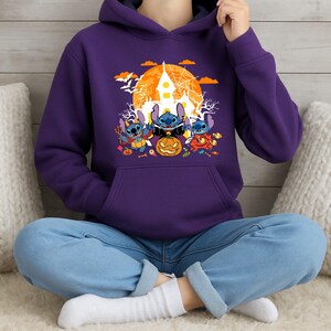 May include: A purple hoodie featuring a Halloween-themed graphic. The graphic includes three cartoon characters dressed in costumes, a jack-o'-lantern, a haunted house, and a full moon. The hoodie has a kangaroo pocket and a drawstring hood.
