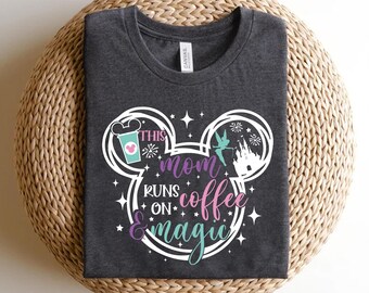 May include: A dark gray t-shirt with a white Mickey Mouse silhouette. The silhouette includes a coffee cup, Tinkerbell, and Cinderella's castle. The text on the shirt reads "This Mom Runs on Coffee & Magic".