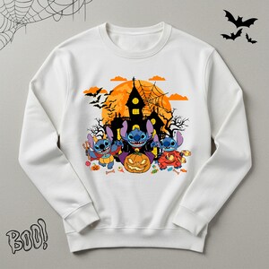 May include: White sweatshirt featuring a Halloween-themed graphic. The design includes three blue cartoon characters dressed in costumes, a jack-o'-lantern, a haunted house, bats, and the word "Boo!".