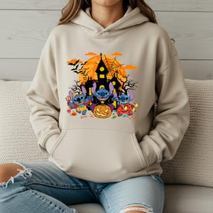 May include: Beige hoodie featuring a Halloween-themed graphic. The design includes three blue cartoon characters, a jack-o'-lantern, a haunted house, bats, and a full moon. The hoodie has a kangaroo pocket and a drawstring hood.