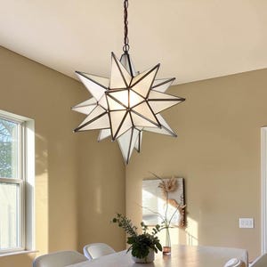 May include: A multi-pointed star-shaped pendant light fixture featuring translucent white glass panels framed in dark metal. The geometric lantern hangs from a dark chain and emits a soft, warm glow from its center, illuminating the surrounding space.