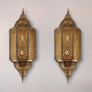 Op de afbeelding: Twee sierlijke, goudkleurige wandkandelaars met ingewikkelde uitgesneden patronen. De kandelaars hebben een lantaarnachtig ontwerp, met een puntige bovenkant en een decoratieve basis. Het licht schijnt door de uitsparingen en creëert een warme gloed.