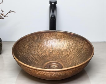 Op de afbeelding: Een gehamerde koperen wastafel met een zwarte kraan. De wastafel heeft een getextureerd oppervlak en een bronzen afwerking. De kraan is zwart en heeft een modern design. De wastafel is ovaal van vorm en staat op een lichtgekleurd aanrecht.