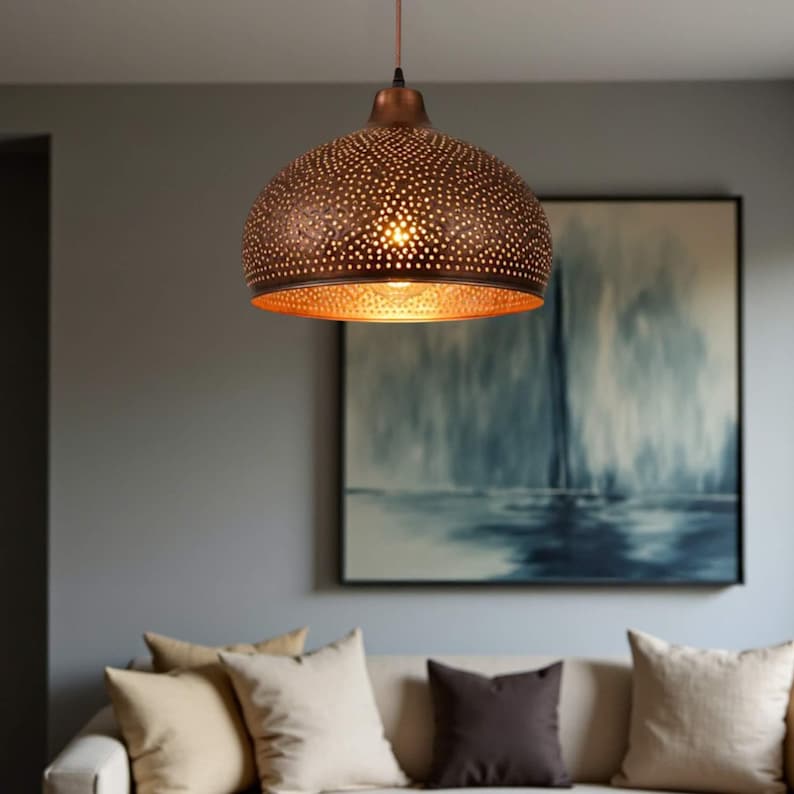 May include: A bronze-colored pendant light fixture with a perforated dome-shaped shade. The light fixture hangs from a thin cord and illuminates the interior with a warm glow. The fixture is suspended above a sofa with neutral-colored pillows.