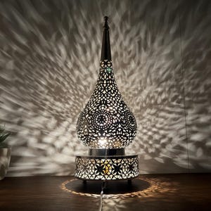 May include: A silver table lamp with a decorative, intricate, cut-out design. The lamp has a round base and a tall, pointed top. The lamp is lit and casting a pattern of light on the surface below.