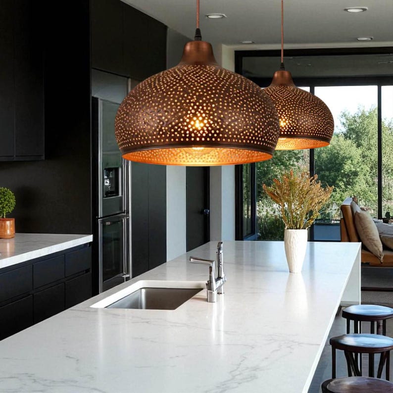 May include: Two dome-shaped pendant lights with a dark bronze exterior finish and intricate laser-cut perforations. The interior of the lamps features a warm, reflective gold-toned surface that glows when illuminated, suspended by thin cords.