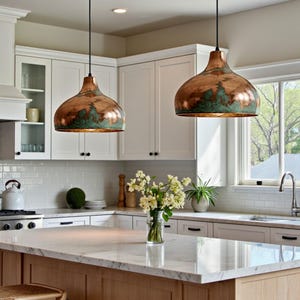 May include: Two copper pendant lights hang above a kitchen island. The lights have a dome shape with a brown and green patina. The kitchen features white cabinets, a marble countertop, and a window.