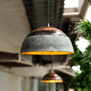 May include: Dome-shaped pendant light with a textured, weathered appearance. The upper portion is a dark, reddish-brown, transitioning to a greenish-gray midsection. The interior glows with a warm, golden light. A second light is visible in the background.