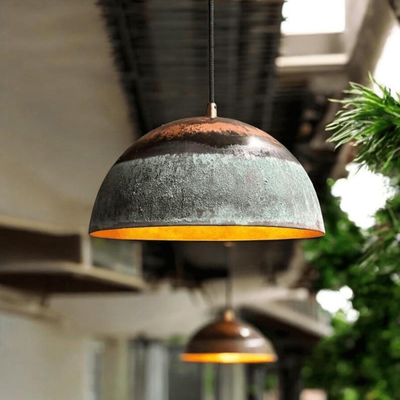 May include: Dome-shaped pendant light with a textured, weathered appearance. The upper portion is a dark, reddish-brown, transitioning to a greenish-gray midsection. The interior glows with a warm, golden light. A second light is visible in the background.
