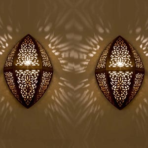 May include: Two gold metal wall sconces with intricate cut-out designs. The sconces are illuminated from within, casting a warm glow.