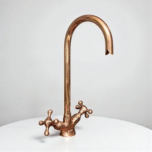 Könnte beinhalten: Ein kupferfarbener Küchenhahn mit gebogenem Auslauf und kreuzförmigen Griffen. Der Wasserhahn ist auf einer weißen Oberfläche montiert und hat ein Vintage-Design. Die Gesamthöhe des Wasserhahns beträgt ca. 38 cm.