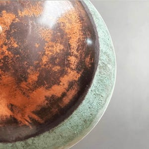 May include: Close-up of a ceramic bowl with a distressed, aged appearance. The interior features a mottled pattern of brown and orange hues, while the rim has a textured, greenish-blue finish. The bowl's surface reflects light, highlighting its unique texture.