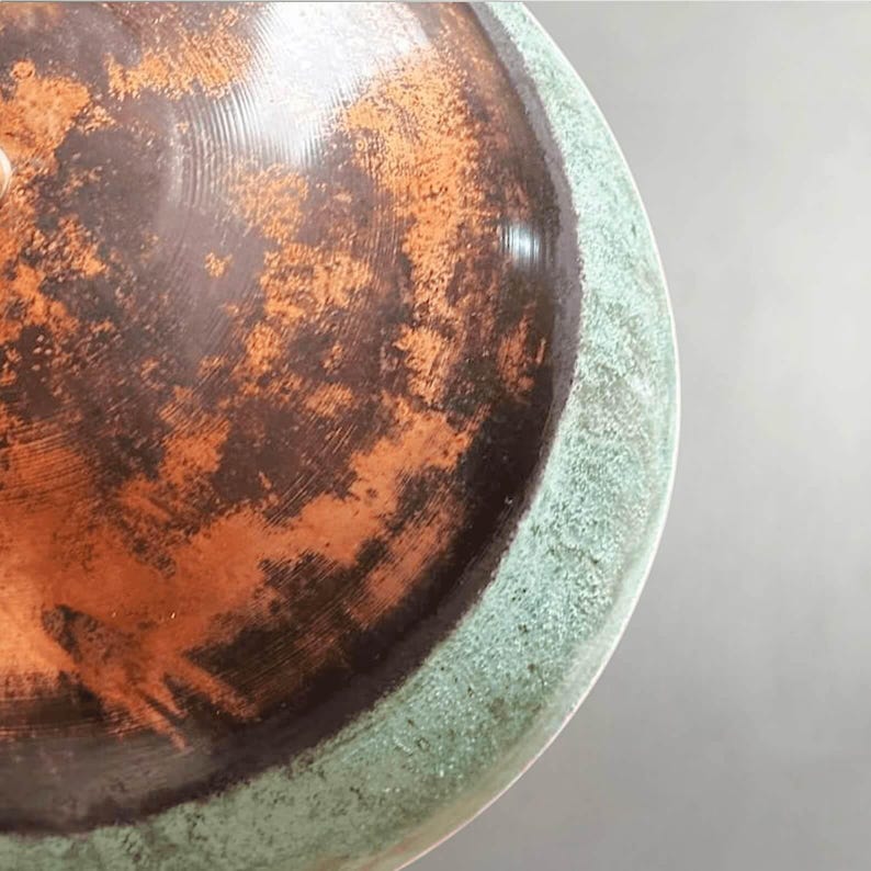 May include: Close-up of a ceramic bowl with a distressed, aged appearance. The interior features a mottled pattern of brown and orange hues, while the rim has a textured, greenish-blue finish. The bowl's surface reflects light, highlighting its unique texture.