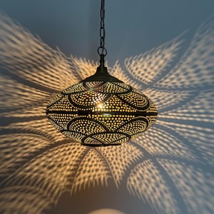 Puede incluir: Una lámpara colgante de latón antiguo con un diseño estampado. La luz está suspendida de una cadena y proyecta intrincadas sombras en la pared. La lámpara tiene un brillo cálido y acogedor.