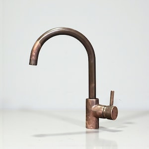 May include: A copper kitchen tap with a curved spout and a single lever handle. The tap has a cylindrical base and a simple, modern design. The finish has a slightly aged appearance.