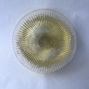 May include: A clear glass plate with a radial ribbed design. The center of the plate has a yellow tint, and the ribbed pattern extends outwards. The plate is circular and appears to be made of glass.