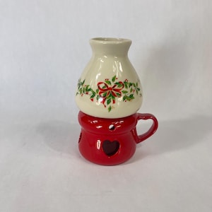 May include: A two-piece ceramic candle warmer. The top piece is cream-coloured with a holly and bow design. The bottom piece is red with heart-shaped cutouts and a handle. A festive holiday decoration.