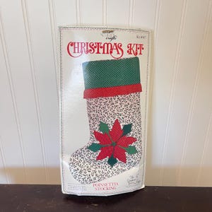 Puede incluir: Un kit de Navidad con un calcetín de flor de Pascua. El calcetín tiene un fondo blanco con un pequeño estampado floral. La parte superior es verde con lunares blancos, y una banda roja la separa del cuerpo principal. Un diseño de flor de Pascua está en el frente.