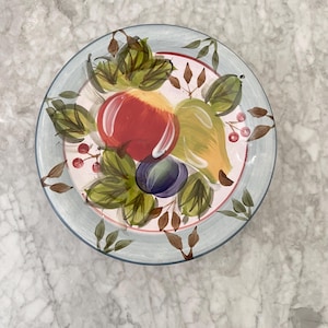 Puede incluir: Plato de cerámica redondo pintado a mano, con un borde azul claro y un diseño central de frutas y hojas coloridas. La fruta incluye una manzana roja, una pera verde y una ciruela morada, rodeadas de hojas verdes y pequeñas bayas rojas.