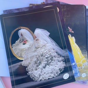 May include: A collection of vintage wedding dress pattern books. The main book shows a bride in a white lace wedding dress and veil, reflected in a gold-framed mirror. Other books display crochet dress designs.