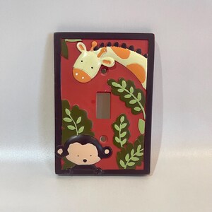 May include: A decorative light switch cover with a jungle animal theme. The cover has a red background with a giraffe, monkey, and green leaves. The giraffe is yellow and orange, and the monkey is brown. The border is brown.