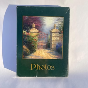 Puede incluir: Un álbum de fotos verde oscuro con la palabra "Photos" en letras doradas. La portada del álbum presenta una pintura de una puerta de jardín con pilares de piedra, flores rojas y un camino.