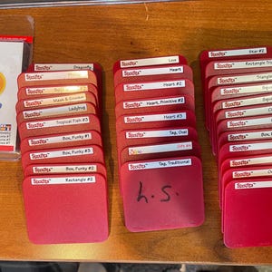 Puede incluir: Montones de tarjetas rojas troqueladas Sizzix con varias formas y etiquetas, incluyendo "Dragonfly" y "Rectangle #2". Un paquete de troqueles Sizzix para manualidades es visible a la izquierda. Las tarjetas están organizadas en tres montones.